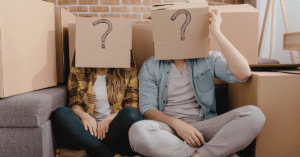 Two people sitting down in front of moving boxes and wearing a box with a question mark on their heads.