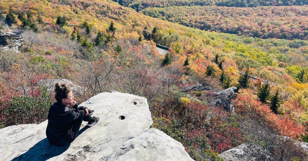 Woman sitting on a rock overlooking trees with fall colors after a hike.