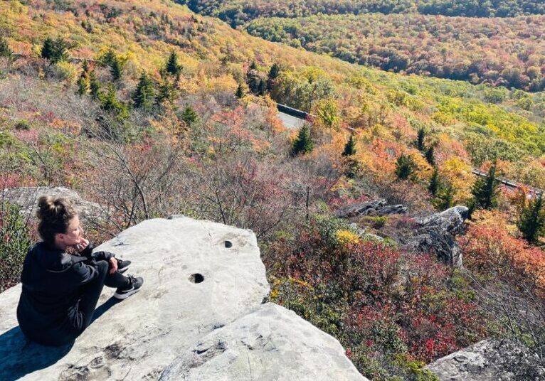 Woman sitting on a rock overlooking trees with fall colors after a hike.
