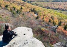 Woman sitting on a rock overlooking trees with fall colors after a hike.