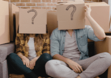 Two people sitting down in front of moving boxes and wearing a box with a question mark on their heads.